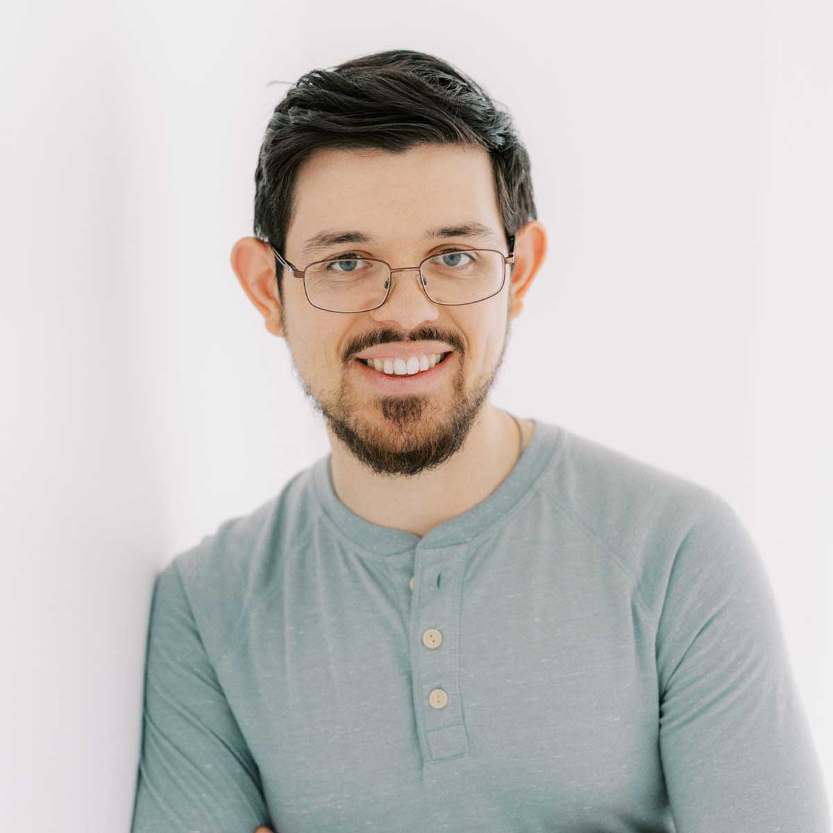 artist Adam Hansen smiles as he leans onto a white wall in a sage green shirt. he has black hair , a beard and wears glasses